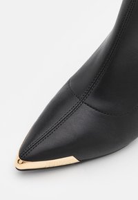 Bottine en cuir noir avec un bout pointu, dotée d'une quincaillerie dorée à la pointe et de coutures décoratives le long des côtés.