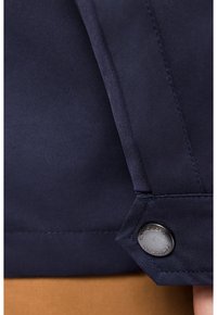 Morska niebieska rękaw kurtki z gładkiego materiału, guzik z wygrawerowanym napisem oraz kontrastującym jasnobrązowym materiałem pod spodem.