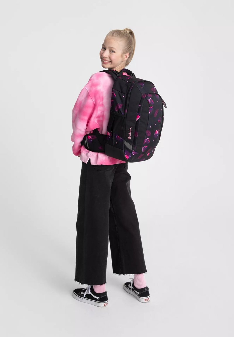 Une adolescente souriante se retournant, vêtue d'un sweatshirt rose tie-dye, de pantalons noirs, de baskets noires et portant un sac à dos noir avec un imprimé floral rose.