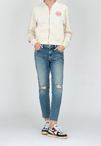 Jasno beżowa bomberka zapinana na zamek z czerwonym logo, zestawiona z podartymi niebieskimi dżinsami oraz czarno-białymi trampkami z czerwonymi akcentami.