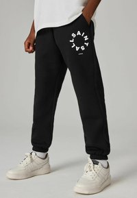 Sorte sweatpants med hvid logo-print, med sidelommer og ankelmanchetter. Parret med hvide sneakers, der viser et struktureret stofdesign.