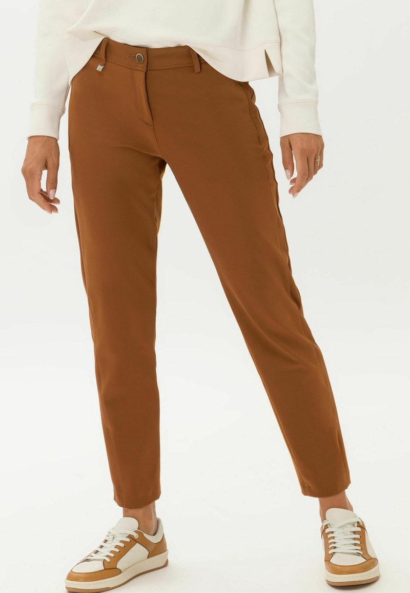 Pantalon marron coupe slim avec bouton et fermeture éclair à l'avant, porté avec des baskets blanches et marron et un haut blanc à manches longues.