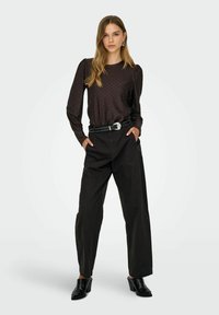 Schwarzes Langarmoberteil mit strukturiertem Muster, kombiniert mit hochgeschnittenen schwarzen Hosen. Model trägt schwarze hohe Schuhe und einen Nieten-Gürtel.
