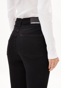 Jeans noirs taille haute avec une texture lisse, présentant une étiquette patch à l'arrière et deux poches arrière, associés à un haut blanc à manches longues.