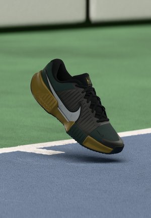 Baskets Nike noires, vertes et marron flottant au-dessus d'une surface de terrain de tennis bleue et verte près d'une ligne blanche.