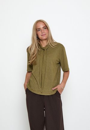 Lyshåret kvinde med langt hår iført en olivengrøn kortærmet top med nakkebånd og mørke, brede bukser med smalle striber.