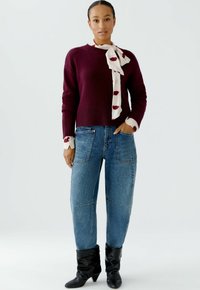 Bordeaux gebreide trui met geribbelde zoom, gecombineerd met lichtblauwe baggy jeans met opgestikte zakken, en zwarte puntige enkelboots. Witte sjaal met rode lippenprint.