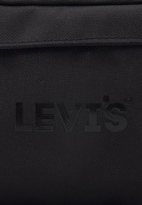 Mustaan kangasta, jossa on tekstuuri, ja jossa on kohokuvioitu "LEVI'S"-logo kiiltävässä pinnassa, sijoitettuna keskelle hyvin näkyvästi.