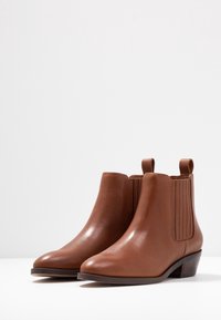 Botas de piel marrón hasta el tobillo con puntera redondeada, paneles laterales elásticos y tacón bajo apilado. Textura lisa con diseño minimalista.