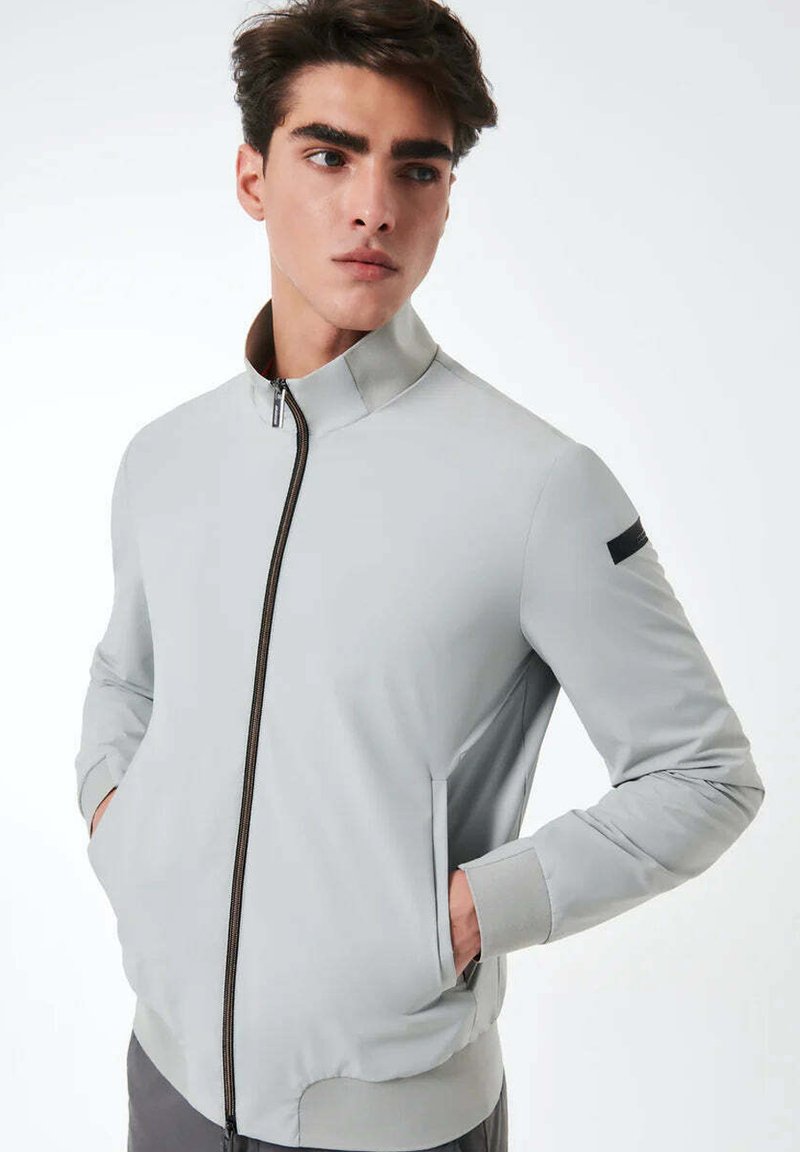 Giacca grigio chiaro con colletto alto, zip frontale e tasche laterali; presenta una texture liscia e un accento nero sulla spalla.