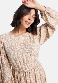 Robe beige à motifs floraux avec une taille cintrée, de longues manches bouffantes et un col rond. Fabriquée en tissu léger.