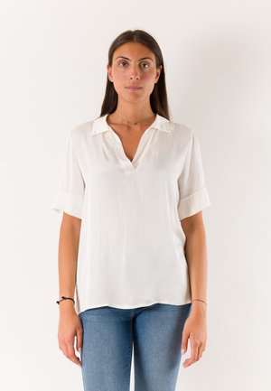 Donna con capelli lunghi e scuri indossa una camicetta bianca a maniche corte e jeans blu, in piedi davanti a uno sfondo chiaro e uniforme, rivolta in avanti.