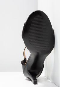 Sandale noire à talon haut avec semelle texturée, talon fin et bride à la cheville, positionnée semelle vers le haut contre un fond blanc d’angle.