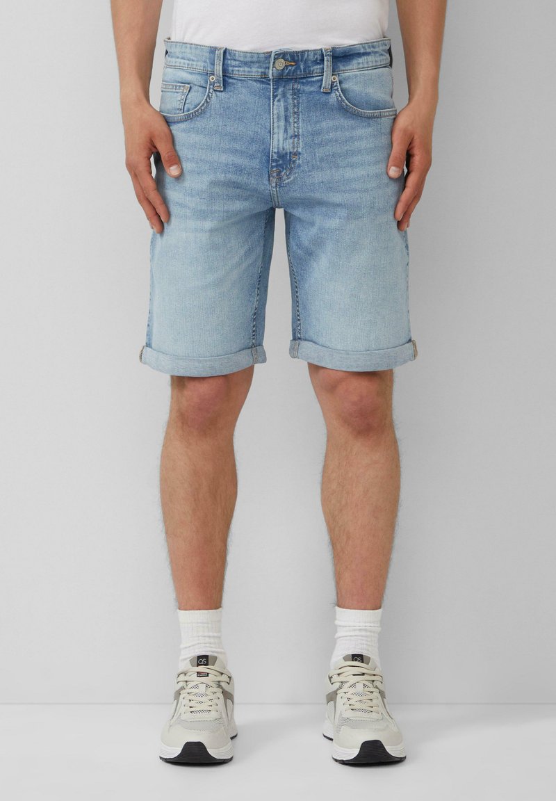 Shorts en jean bleu clair avec des ourlets retroussés, cinq poches et une fermeture à bouton à l'avant. Portés avec des chaussettes blanches et des baskets claires.