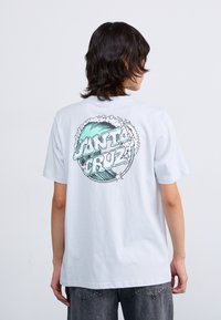 T-shirt bianca in cotone con una grande grafica sul retro con "Santa Cruz" in teal e nero, circondata da design di onde e nuvole.