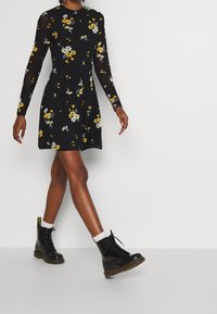 Vestido negro de manga larga con patrones florales coloridos, cintura entallada, falda acampanada, combinado con botas negras con cordones y calcetines blancos hasta el tobillo.