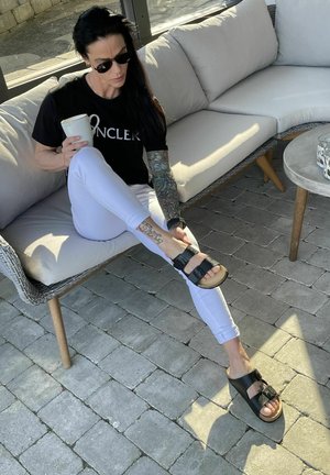 Žena s tetovaniami a slnečnými okuliarmi sedí na vonkajšej pohovke, drží hrnček a upravuje si sandál na nohe, slnečná kamenná terasa.