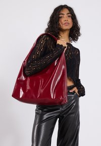 Borsa a spalla bordeaux lucida realizzata in pelle liscia, caratterizzata da una forma morbida e rilassata. Abbinata a un top nero all'uncinetto e pantaloni in pelle.