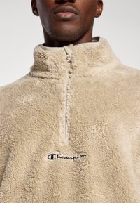 Béžový fleecový pulóver s golierom na zips, s jemnou textúrou a čiernym vyšívaným logom "Champion" na prednej strane.