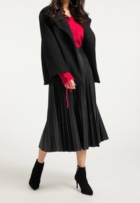 Manteau noir oversize porté sur un pull en maille rouge aux manches texturées, associé à une jupe plissée noire et des bottines à talons noires.