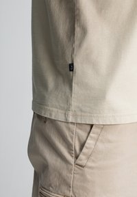 Close-up van beige katoenen broeken met een zijzak en de onderkant van een lichtbeige shirt met een klein zwart merklabel aan de linkerkant.