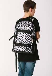 Zaino a scacchi bianco e nero con più scomparti con cerniera, spallacci regolabili e un accessorio a forma di portachiavi attaccato.