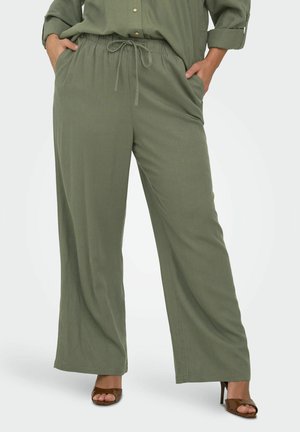 Kvinne iført løstsittende olivengrønne bukser med snøring og sidelommer, kombinert med brune høyhælte sandaler med åpne tær.