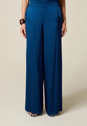 Donna che indossa pantaloni a vita alta a gamba larga blu navy con tasche laterali e sandali turchesi con punta aperta su uno sfondo semplice.