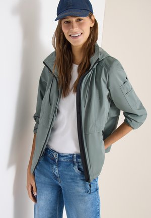 Mujer sonriente con una chaqueta verde claro con cremallera, camiseta blanca, vaqueros azules y una gorra de béisbol azul oscuro, de pie con una mano en el bolsillo.