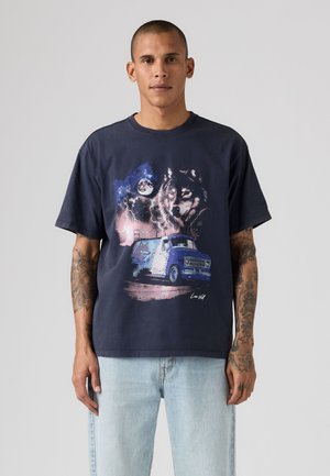 Mand iført en navyblå t-shirt med grafik af en ulv, måne, stormfuld himmel og blå varevogn, parret med lyseblå jeans, stående mod en grå baggrund.