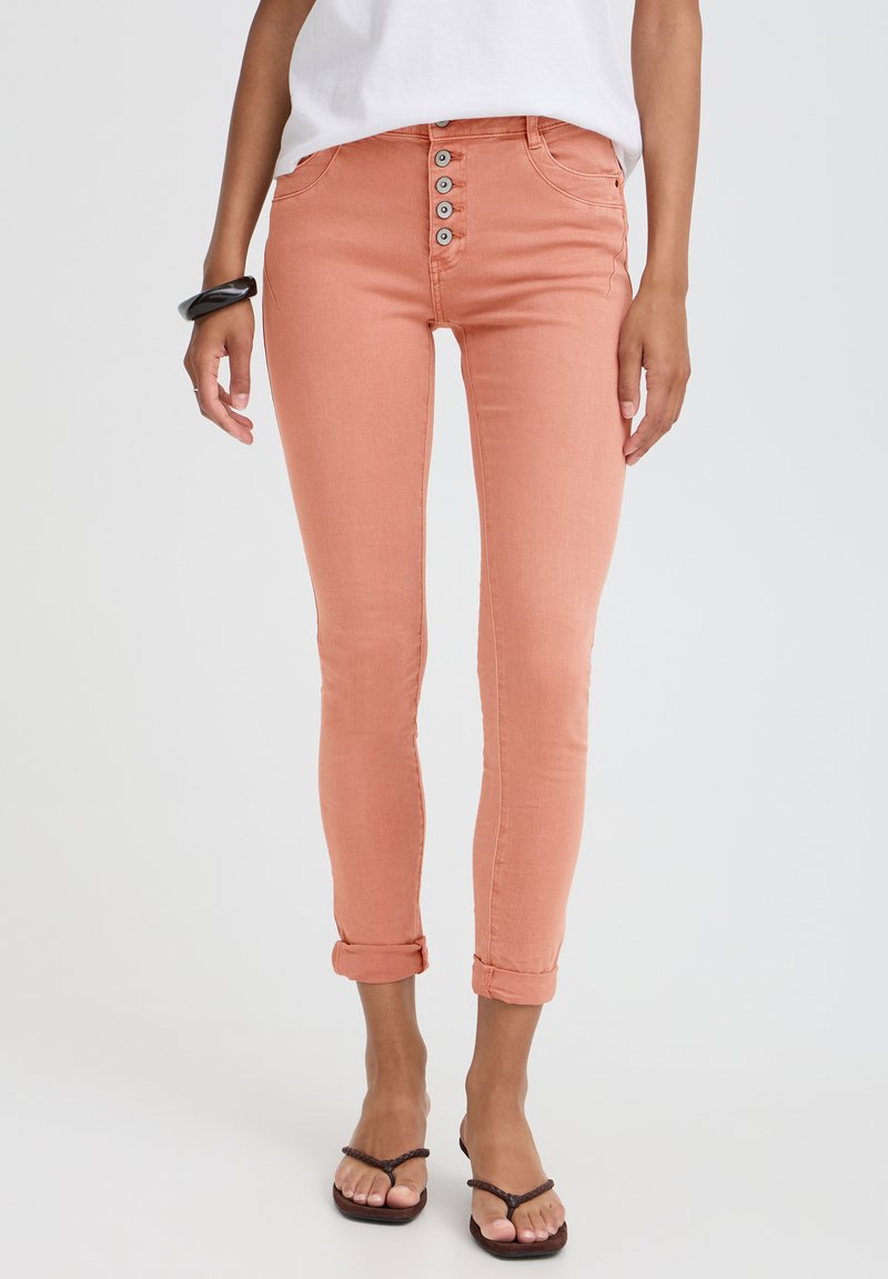 Personne portant un jean skinny couleur pêche avec cinq boutons apparents, un haut blanc, un bracelet noir et des sandales, debout devant un fond clair et uni.