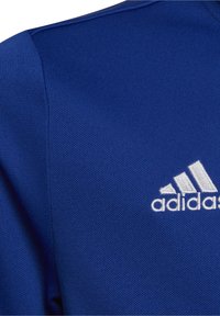 Tela texturizada azul con el logo bordado de Adidas en blanco, que presenta tres franjas y el nombre de la marca en la parte superior de la prenda.