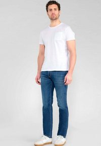 T-shirt en coton blanc avec une poche et un détail de logo, associé à un jean en denim bleu et des baskets blanches avec une semelle beige.