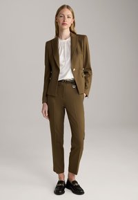 Brauner maßgeschneiderter Anzug mit einem zweireihigen Jacket, weißer plissee Bluse und schwarzen Slippern mit goldenen Hardware-Akzenten.