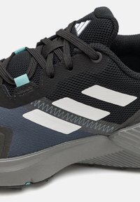 Black athletic shoe featuring a mesh upper, turquoise lace accent, and three white stripes. Textured gray sole with pronounced grooves.