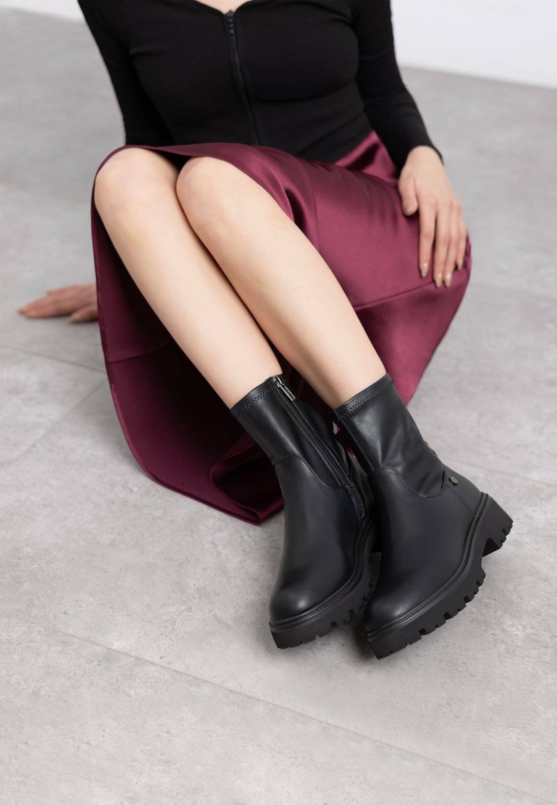 Botas de tobillo negras de cuero, con una suela de goma gruesa, cremallera lateral y un diseño elegante. Llevadas con una falda de satén burdeos.