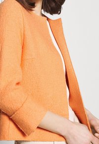 Veste texturée orange avec un col rond, des manches trois-quarts, et une coupe ouverte sur le devant, associée à un haut de couleur claire en dessous.