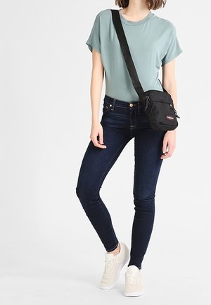 T-shirt bleu clair, skinny jeans noirs et baskets beiges, assortis à un petit sac crossbody noir, se tenant devant un fond uni.