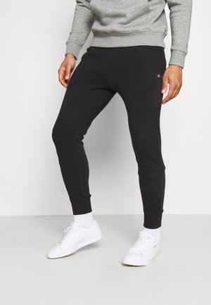 Homme portant un pantalon de jogging noir ajusté avec un petit logo blanc « ck » sur la cuisse, des baskets blanches et un sweat gris, debout sur un sol clair.
