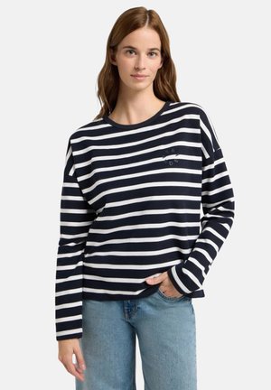 Young woman wearing a navy and white striped long-sleeve shirt and light blue jeans, standing with one hand in pocket.