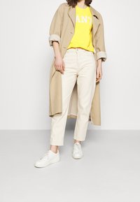 Béžový lehký kabát, žluté tričko s bílým potiskem, krémové kalhoty a bílé tenisky. Tento outfit předvádí neformální, vrstvený styl.