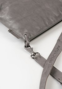 Graue Lederhandtasche mit glatter Textur, ausgestattet mit einem Reißverschluss, verstellbarem Schulterriemen und metallischen Beschlägen mit einem Verschluss und Logodetail.