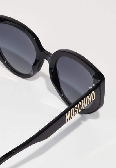 Juodos saulės akinės su per didelėmis kampuotomis rėmeliais, tamsintais objektyvais ir auksiniu "MOSCHINO" logotipu ant matinio juodo šventyklos.
