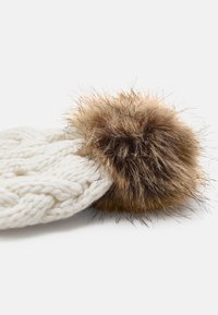 Bonnet en tricot blanc avec un grand pompon rond en fausse fourrure marron. La texture est épaisse avec un motif tressé, mettant en avant la chaleur.