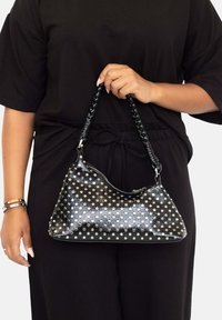 Bolso de cuero negro con lunares plateados, forma curvada y asa trenzada. Presenta un cierre con cremallera y textura suave.