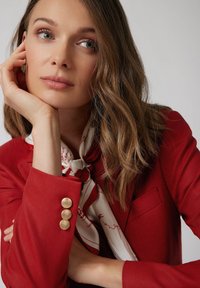 Czerwony żakiet o teksturowanej powierzchni z trzema złotymi guzikami na rękawie oraz kremowym wzorzystym szalem. Dekolt ma zawiązany wzór w czerwone paski.