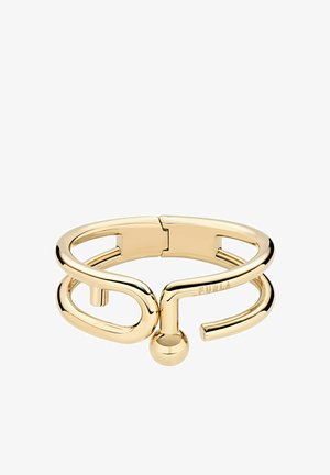 Zelta toņa metāla aproce ar minimālistisku dizainu, kurā ir izliekts formu elements un sfērisks akcenta elements. Zīmola nosaukums iestrādāts aproces iekšpusē.