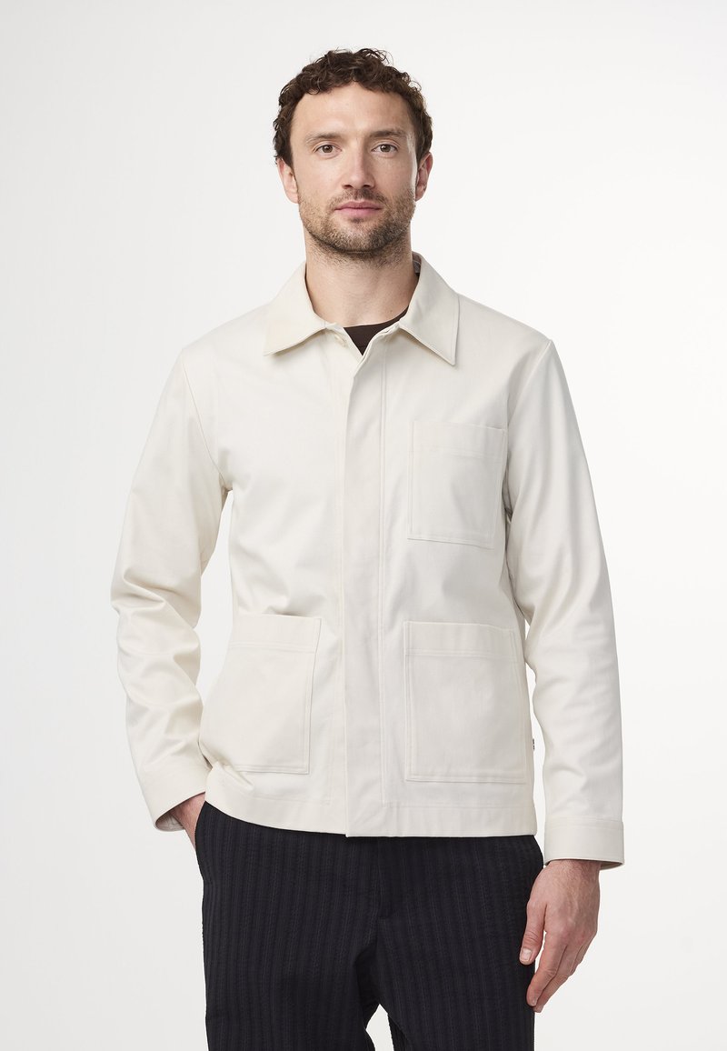 Ljusbeige jacka gjord av bomullsliknande material, med en spredd krage, knappslutning fram och två stora påsydda fickor. Svarta texturerade byxor.