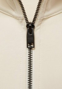 Giacca a zip color crema con una cerniera nera, caratterizzata da un tessuto testurizzato e un design minimalista e liscio. Accentuata da un tirazip con il logo.