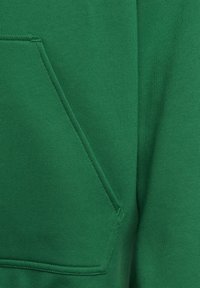 Primo piano di un tessuto felpa verde con tasca a marsupio, texture liscia e cuciture rinforzate lungo i bordi.